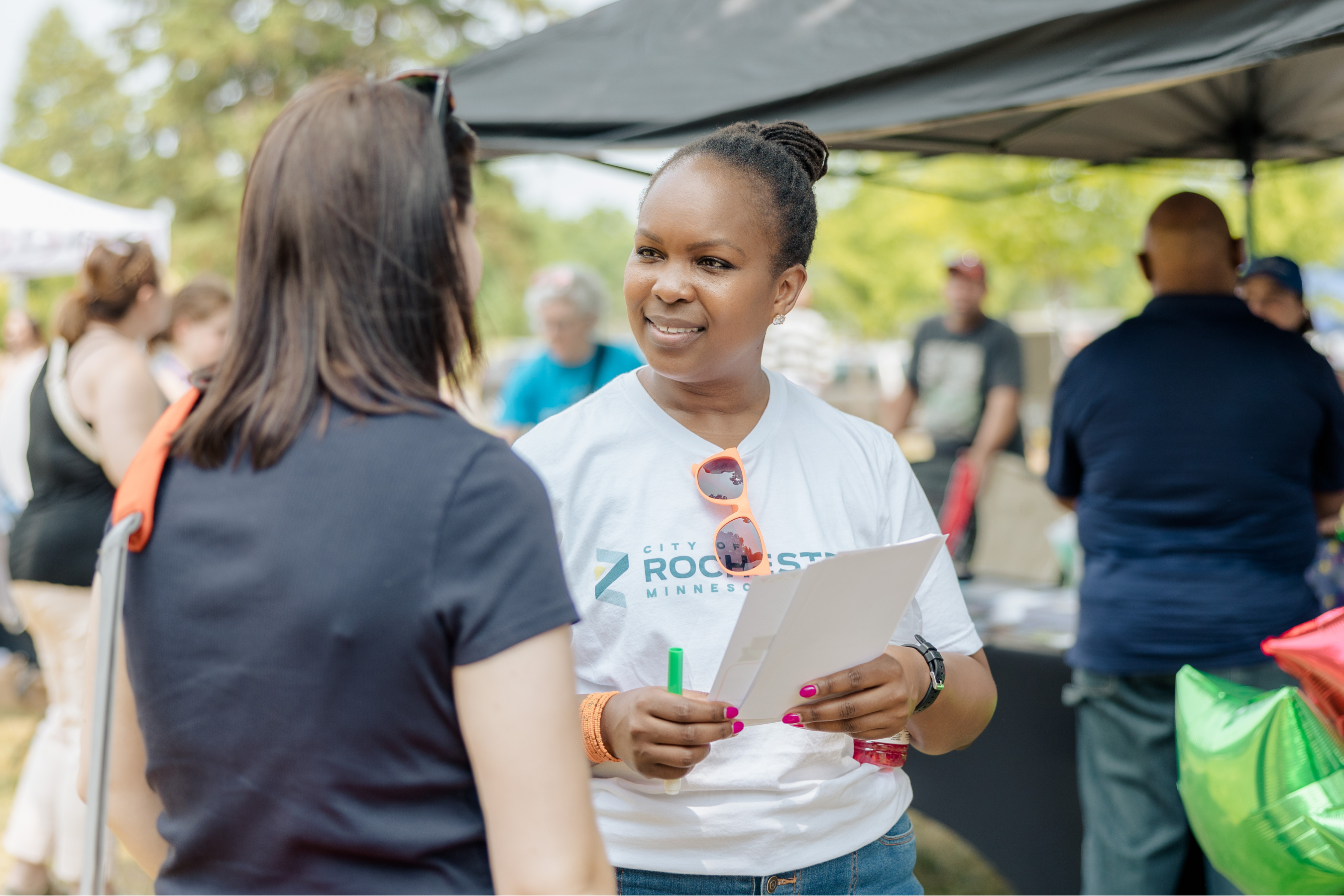 two women conversing at a community event with tents and people gathered around